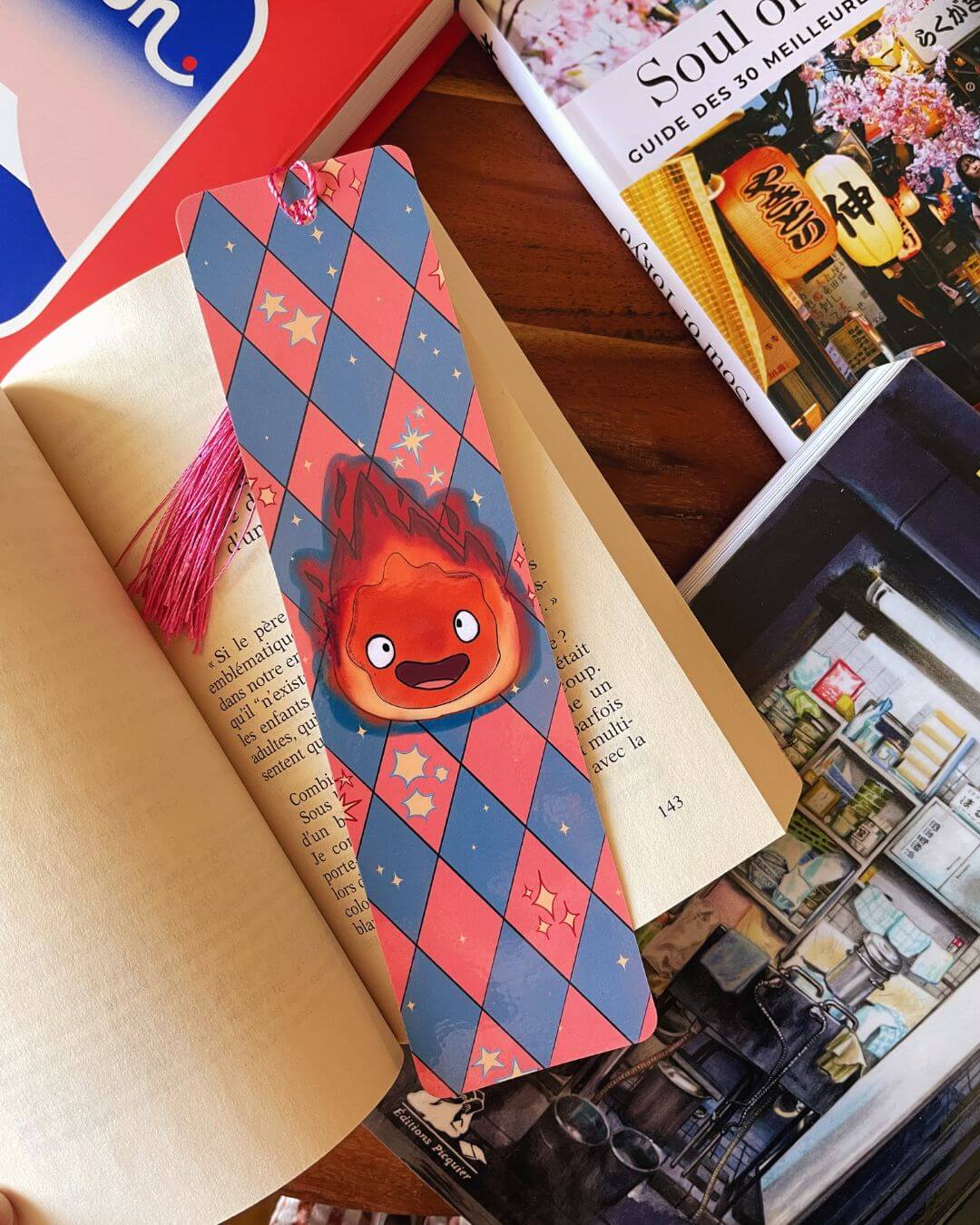 marque-page illustré avec le personnage Calcifer sur un fond à losanges bleus et roses, avec un pompon rose