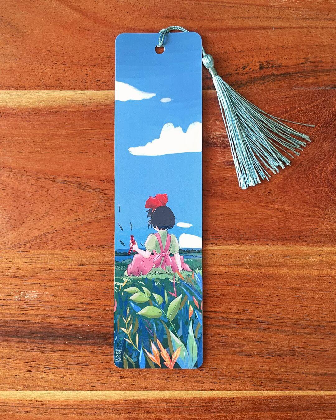 marque-page illustré représentant Kiki la petite sorcière de dos dans un champ de fleurs, avec un pompon bleu clair,