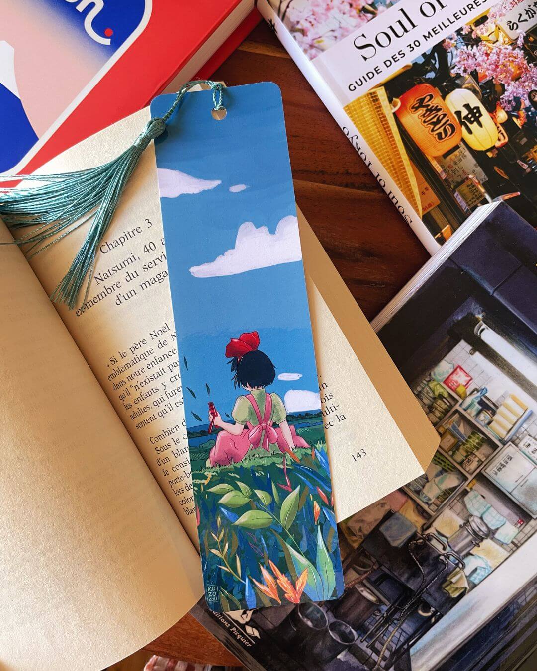 marque-page illustré représentant Kiki la petite sorcière de dos dans un champ de fleurs, avec un pompon bleu clair,