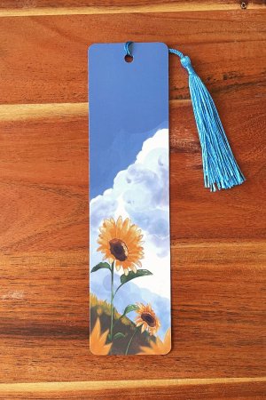 marque-page illustré représentant un champ de tournesols sous un ciel bleu nuageux, avec un pompon bleu vif,