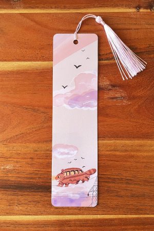 Photo d'un marque-page illustré représentant le Chat Bus courant sur des fils électriques sous un ciel rose et violet, avec un pompon rose clair