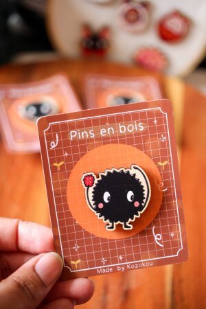 Photo d'un pin's en bois d'érable représentant une noiraude, posé sur un tissu, montrant le grain naturel du bois.
